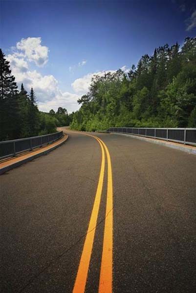 A double yellow centre line running down a sealed road between trees under a blue sky