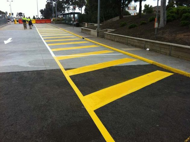 Bright yellow hatched pedestrian crossing bars painted across a car park entrance, workers in the distance