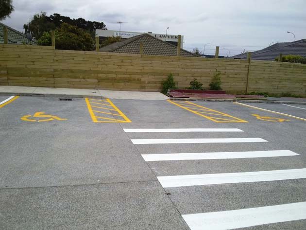 Two mobility parking bays marked in yellow with wheelchair symbols, white crossing stripes in front and a timber fence behind