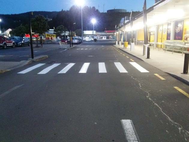 A white striped pedestrian crossing outside a row of shops at dusk, the car park lit by lamps