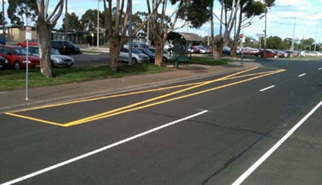 Yellow hatched median markings on a road beside parked cars under tall trees