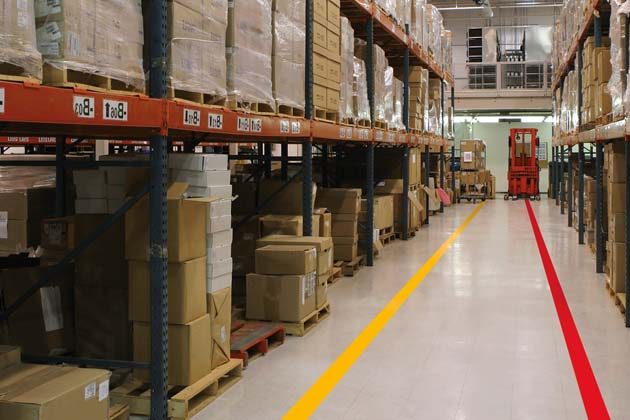 A warehouse aisle between racks of boxed stock with a yellow line and a red line painted along the floor