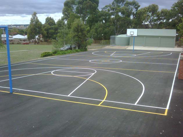 A school court marked with white basketball lines and yellow netball lines, a hoop and blue posts at one end